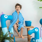 Child wrapped in a blue striped towel sitting on a chair with blue dragon-shaped chairs and a bowl in the background.